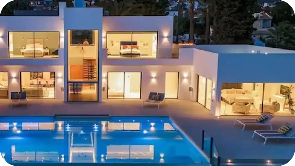 Een grote, verlichte designvilla in de avond met enorme glazen puien die inkijk geven in de sfeervolle slaapkamers en woonruimtes, grenzend aan een weerspiegelend zwembad