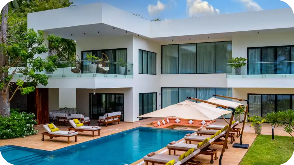 Een luxe witte villa met twee verdiepingen in een tropische stijl, voorzien van een groot zwembad, ligstoelen en beige parasols in de tuin
