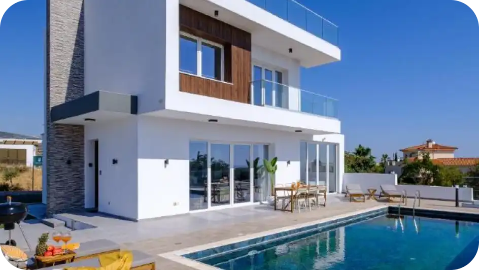 Een moderne witte villa met twee verdiepingen, houten accenten en glazen balkons, met een strak zwembad en een eettafel op het terras onder een helderblauwe lucht