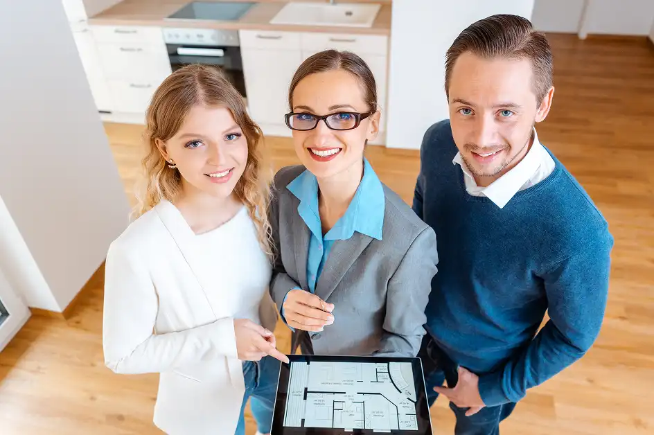 Een vrouwelijke makelaar in een grijs pak toont glimlachend een digitale plattegrond op een tablet aan een jong stel in een nieuwe woning